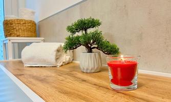 A stylish bathroom with a wooden table, a small plant, and a red candle holder. Towels and a basket complement the relaxing atmosphere.