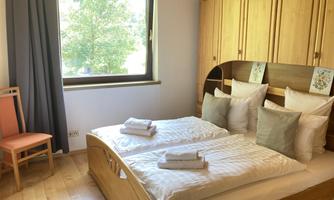 A cozy bedroom with a large double bed and many cushions. A window overlooking a sunny tree outside and a wooden wardrobe complement the space.