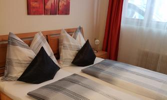 A cozy bedroom with a double bed and decorative cushions. The walls are bright and a window lets in daylight.