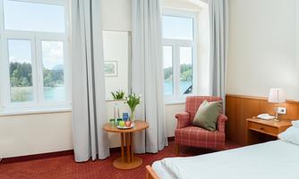 A bright hotel room with a cozy armchair and a small table. Through the windows, there is a view of the landscape and the water.