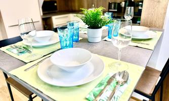 A beautifully set table with white plates, blue glasses, and a small plant in the center. The tablecloth is green, and the surroundings feel modern and inviting.