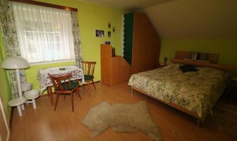 A cozy bedroom with a large bed and a small seating area. The walls are painted in a fresh green color, and the light softly falls through the window.