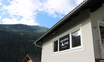 A modern building with a sign that says "Apartments Zula." In the background, majestic mountains and a blue sky are visible.