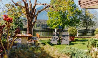 A quiet garden landscape with a large tree and lounge chairs. In the background, the mountains and a wooden fence can be seen.