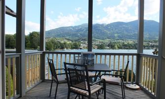 A cozy balcony with a table and chairs overlooking a lake and surrounding mountains. The landscape is green and inviting.