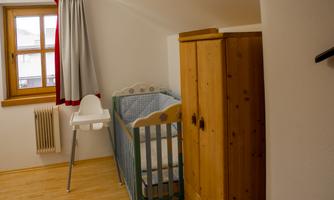 A bright children's room with a crib and a high chair. The room has wooden floors and a window with a red curtain.