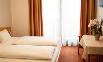 A simple and cozy bedroom with two beds. A table with a flower and large windows lets in plenty of light.