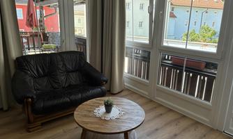 A cozy room with a black leather sofa and a round table. Large windows offer a view of colorful houses and our castle ruins.