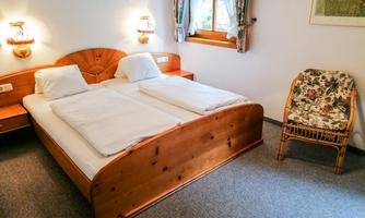 A cozy bedroom with a wooden double bed and two pillows. Next to the bed is a side chair with a floral pattern.