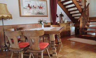 A cozy dining area with a wooden table and chairs in warm colors. In the background, there is a staircase and a decorative wall with a painting.