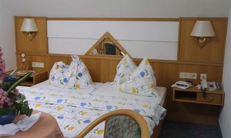 A cozy bedroom with a double bed and colorful cushions. Next to the bed, there is a wooden desk and a small floral decoration on the table.
