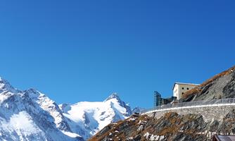An impressive mountain landscape with snow-covered peaks and a clear blue sky. In the foreground, there is a building along a winding path.