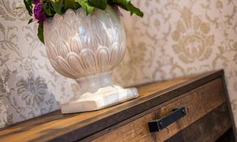 An elegant flower vase stands on a wooden cabinet. The vase is decorated with colorful flowers and placed in front of a stylish wallpaper.