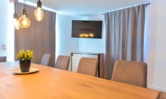 A modern meeting room with a large wooden table and comfortable chairs. A television wall hangs on the wall, and the windows are covered with gray curtains.