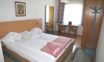 A simple bedroom with a double bed and two pillows. There is a desk, a chair, and a clothes rack, as well as natural light through a large window.