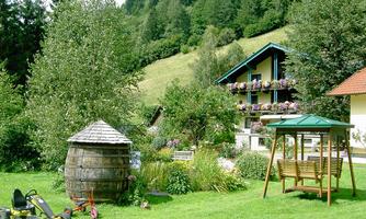 A beautiful garden with a wooden barrel, a swing, and a lovely house in the background. There are green trees and flowers everywhere.
