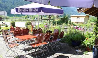 An inviting outdoor area with tables and chairs under a sunshade. Surrounded by green plants and a picturesque landscape.