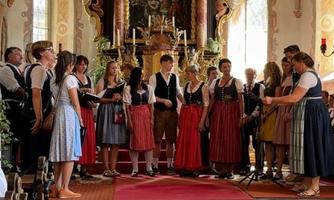 Eine Gruppe von Menschen in traditioneller Tracht steht auf einer Bühne in einer Kirche. Sie scheinen zu singen oder zu proben.