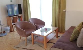 A cozy living room with a sofa and two armchairs. A table is in the middle and a television is visible in the background.