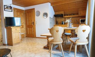 A cozy apartment with wooden decor, a dining table and chairs in heart shapes. In the background, there is a modern kitchen and a television.
