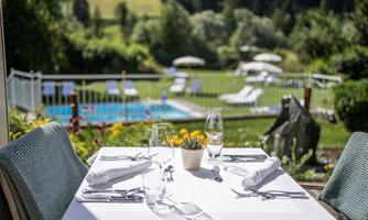 An elegant table set with silver cutlery and a small plant, overlooking a green landscape with a pool and loungers. The area is surrounded by trees and exudes a relaxed atmosphere.