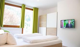 A modern bedroom with a large window and green curtains. A TV displays a news program on the wall.