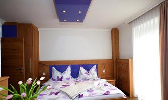 A cozy bedroom with a large bed and colorful pillows. The walls are made of wood, and large windows bring in plenty of natural light.