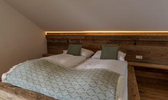 A cozy bedroom with a large wooden bed and green pillows. Warm light and rustic wooden walls create an inviting atmosphere.