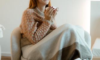 A smiling woman is sitting on a comfortable chair, surrounded by a soft carpet. She is holding a cup in her hand and looks relaxed.