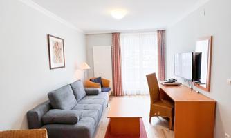 A modern living room with a gray sofa and a wooden table. The room has large windows with curtains and a television on a desk.