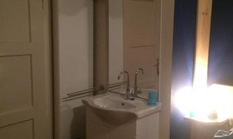 A small room with a sink and a mirror. The walls are painted in neutral colors, and there is a lit area next to the bed.