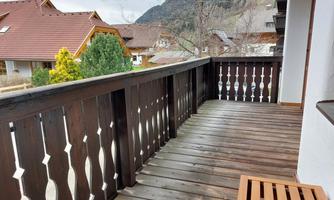 A balcony with a wooden railing and a view of adjacent houses and mountains. The floor is made of wood and it is cloudy.