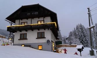 A cozy house in the snow, surrounded by trees. The windows are lit, and there is a calm winter atmosphere in the air.