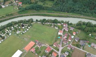 Aerial view of a small village with houses, a soccer field, and a river. Surrounded by plenty of green and trees.