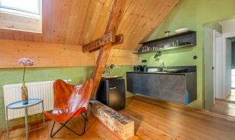 A modern attic apartment with green wall paint and wooden beams. The space features a minimalist kitchenette and a cozy armchair.