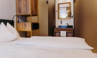 A modern bedroom with a double bed and elegant furniture. In the background, the bathroom with a sink and a mirror can be seen.