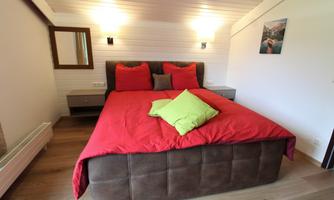 A cozy bedroom with a large, comfortable bed and red bedding. The walls are bright, and there is soft lighting as well as modern decor.