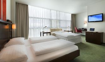 A modern hotel room with two beds, a desk, and a television. Large windows provide plenty of daylight.
