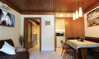 A cozy living room with a dining table and a modern kitchen in the background. A TV is mounted on the wall, and the room is well-lit.