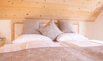 A cozy bedroom with a wooden ceiling and a bed decorated with soft pillows and a blanket. The gentle colors create a relaxed atmosphere.