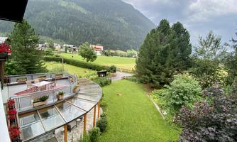 A green garden landscape surrounded by trees and mountains. In the foreground, there is a terrace with flowers.