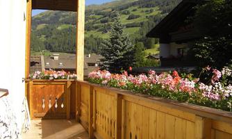 A cozy balcony with a wooden railing and colorful flowers. In the background, green hills and a clear sky can be seen.