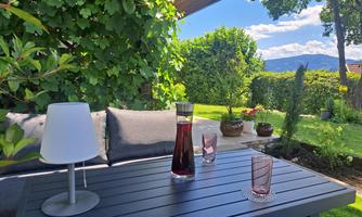 A cozy outdoor area with a table and a beautiful view. On the table are a carafe, glasses, and a lamp, surrounded by lush greenery.