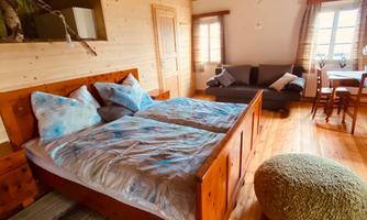 A cozy bedroom with a large bed and wooden furniture. There is a living area with a couch and a table, surrounded by warm, natural light.