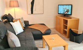 A cozy living room with a gray sofa and a small wooden table. A piece of art hangs on the wall, and a television sits on a sideboard.