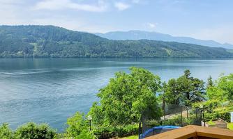 A tranquil landscape with a lake and green trees. In the background, mountains and a clear blue sky can be seen.