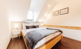 A cozy bedroom with a large bed and wooden details. The walls are light-colored and there are two windows that provide natural light.
