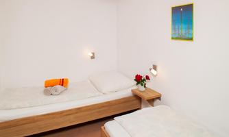 A simple bedroom with two single beds and a bedside table. A small floral arrangement and a towel complete the furnishings.