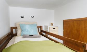 A cozy bed in a small room with a white wall. On the pillow is a blue cover and a green blanket, while decorative eyes are mounted above.