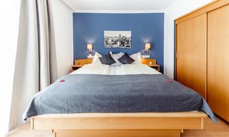 A cozy bedroom with a large bed and blue walls. On the sides, there are night lamps and the curtains are light gray.